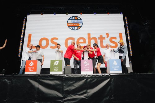 Menschen in roten und weißen Shirts stehen auf Bühne vor großer Leinwand mit Text 'Los geht's!' und Symbolen für Feuer, Erde, Luft und Wasser mit Entfernungen.