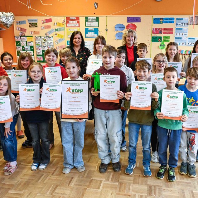 In einem Klassenzimmer steht eine Gruppe von Kindern und zwei Erwachsenen nebeneinander. Die Kinder halten Zertifikate hoch, auf denen "Step" steht, und lächeln in die Kamera. Im Hintergrund sind bunte Wanddekorationen und Lernmaterialien zu sehen. Die Stimmung ist fröhlich und feierlich.
