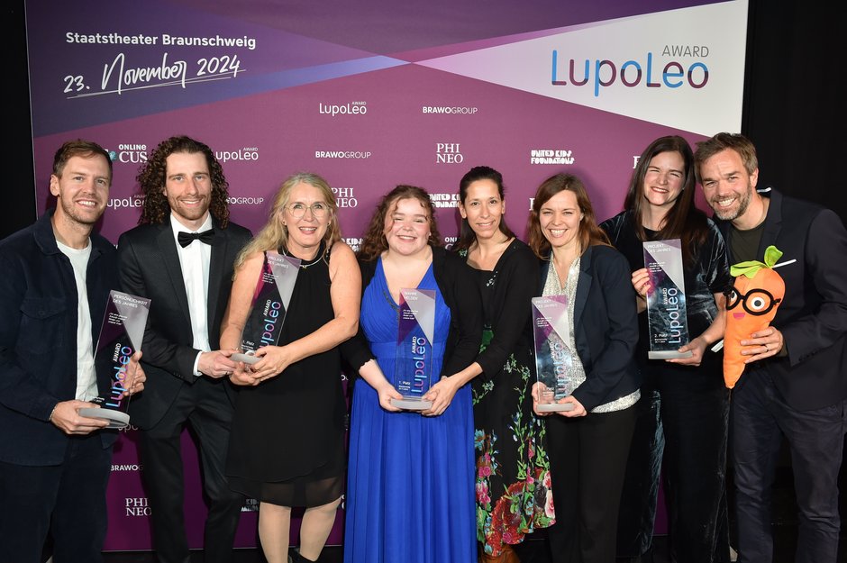 Eine Gruppe von acht Personen posiert lächelnd mit ihren Preisen vor einem farbigen Banner, auf dem "LupoLeo Award" steht. Alle halten ihre Auszeichnungen stolz in den Händen, und einer der Anwesenden hält auch eine Plüschkarotte mit Brille. Die Veranstaltung findet im Staatstheater Braunschweig statt, wie das Banner im Hintergrund angibt. Die Kleidung ist festlich, mit Anzügen und Abendkleidern.
