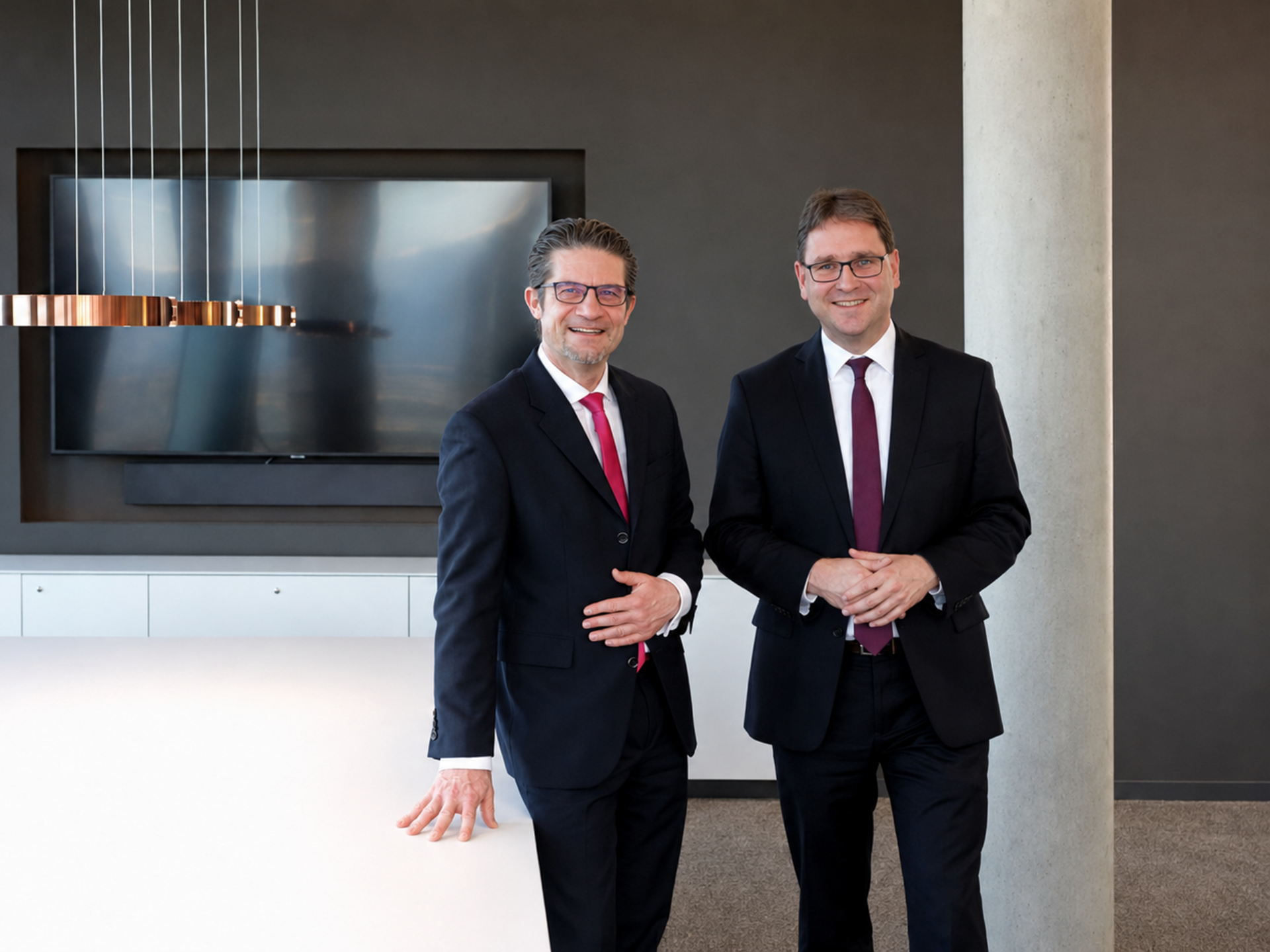 Zwei Männer in Anzügen stehen nebeneinander in einem modern eingerichteten Raum. Beide tragen Brille und Krawatte, der Mann links lehnt mit einer Hand entspannt auf einen weißen Tisch. Im Hintergrund sind ein großer Bildschirm und eine moderne Hängelampe zu sehen. Die Wände und der Boden sind in neutralen Tönen gehalten.