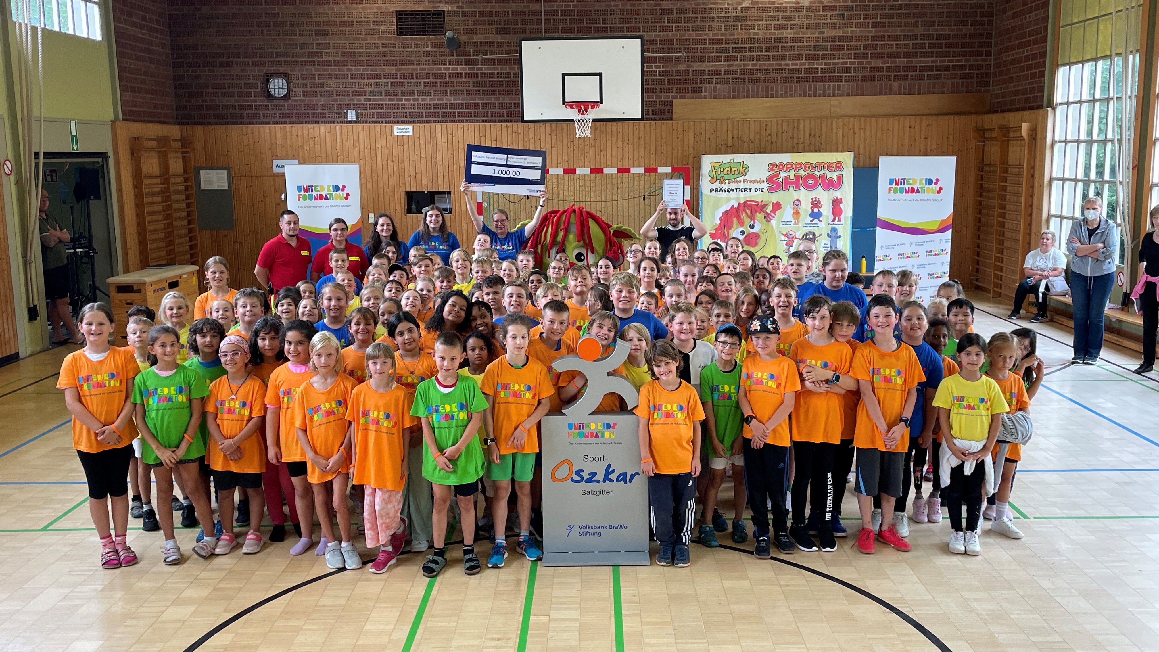 Große Gruppe von Kindern und vereinzelt Erwachsenen steht in bunten Tshirts in einer Turnhalle.